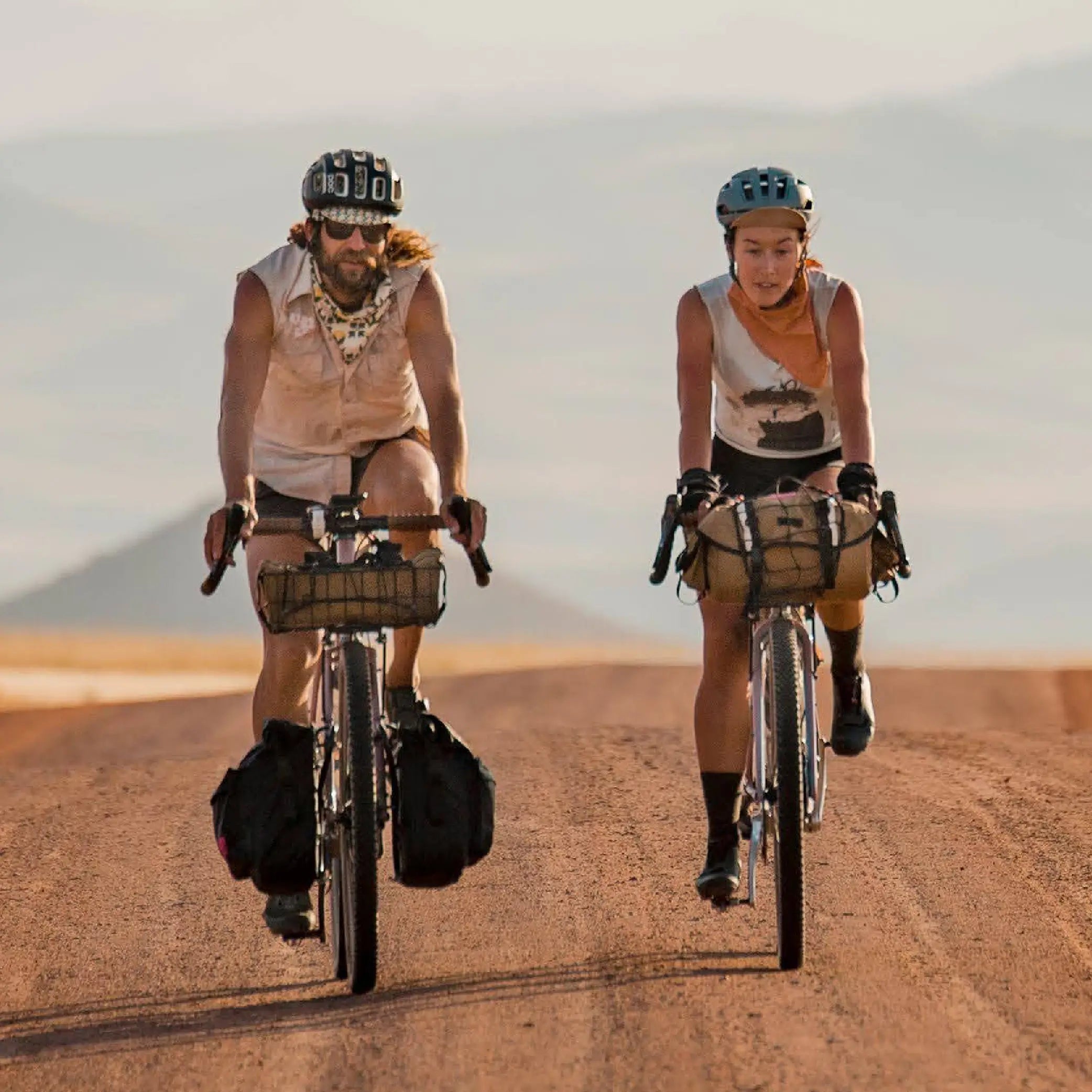 Two people riding bicycles on a desert road with mountains in the background.